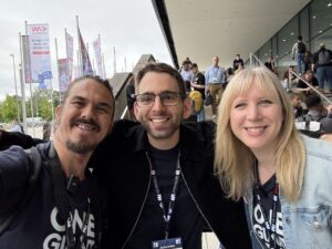 Bart Farrell, an attendee, and Hannah Foxwell smile for a group photo outside the WeAreDevelopers World Congress venue. Attendees gather in the background near branded flags and event signage.